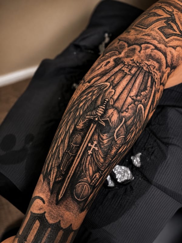 black and grey tattoo of a winged hooded knight resting on a sword under rays of light breaking through clouds on the forearm by HEAVY INK, a tattoo artist in chino, california