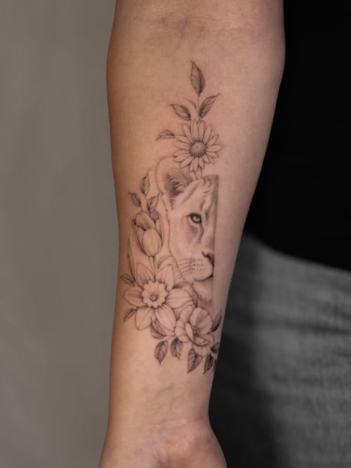 A delicate black and grey tattoo of a lion with flowers on the forearm, featuring fine line details and ornate floral elements — by Melanie Benoit, los angeles