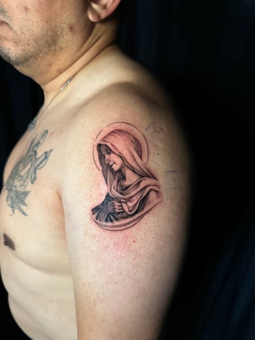 A black and grey realism portrait of the Virgin Mary on the upper arm, featuring detailed shading and a serene expression — by Travis Ross, los angeles