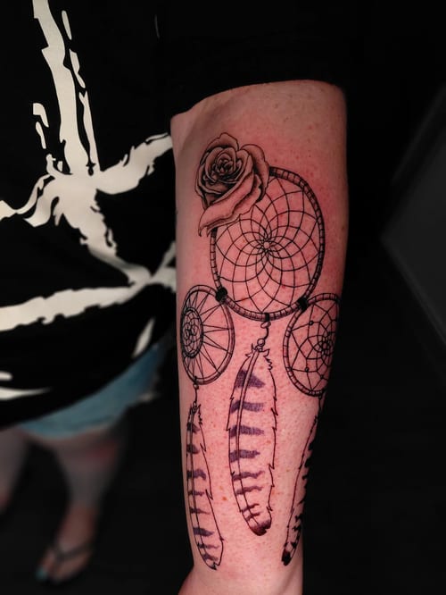 A black and grey linework tattoo of a dreamcatcher with feathers and a rose on the forearm — by anoetic.designs.ultd, los angeles