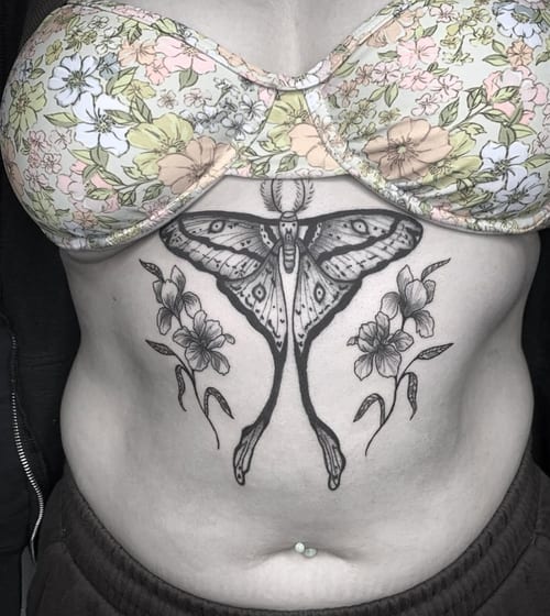 A black and grey ornamental tattoo on the stomach featuring a large butterfly with floral elements on either side — by Shelby Bianco, los angeles