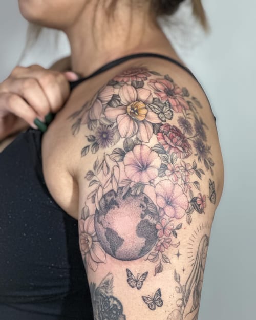A colorful tattoo of the Earth surrounded by flowers and butterflies on the shoulder, blending illustrative and floral styles — by Melanie Benoit, los angeles