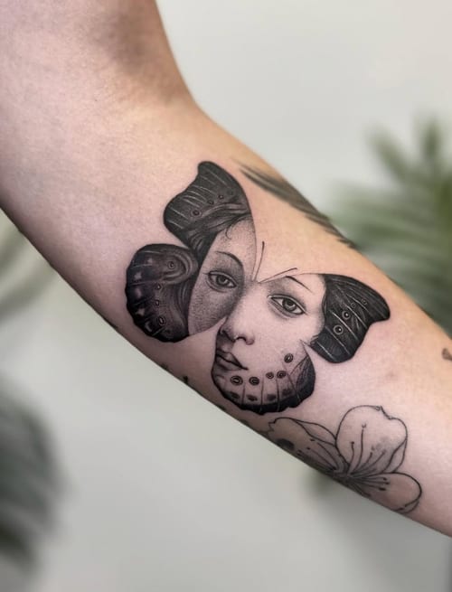 A surreal black and grey tattoo on the inner arm featuring a butterfly with a human face integrated into its wings, showcasing intricate linework and shading — by Jian Liang, los angeles