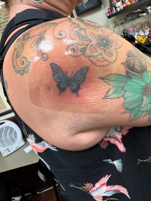 A colorful butterfly tattoo on the shoulder surrounded by floral and ornate designs, done in a traditional style — by Sandra Price, los angeles