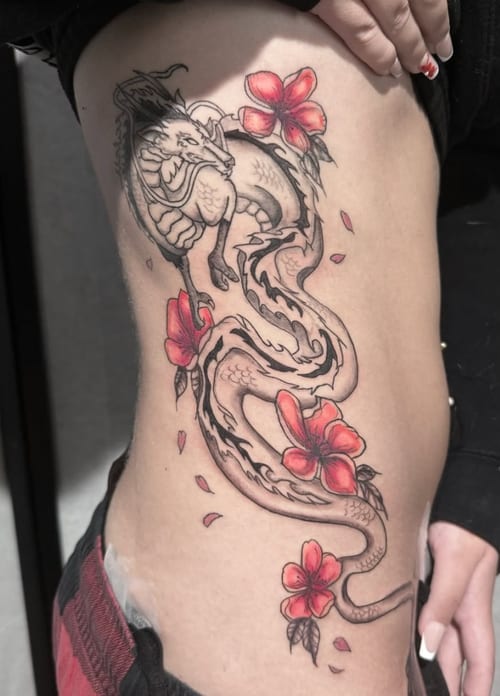 A mixed color tattoo of a Japanese-style dragon intertwined with red flowers on the rib, featuring detailed linework and shading — by Shelby Bianco, los angeles