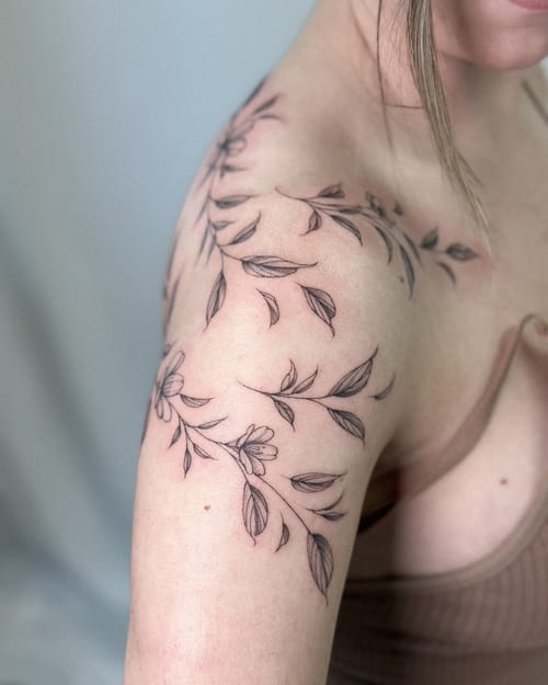 A delicate fine line floral vine tattoo on the shoulder, featuring black and grey ink with intricate leaf and flower details — by Melanie Benoit, los angeles