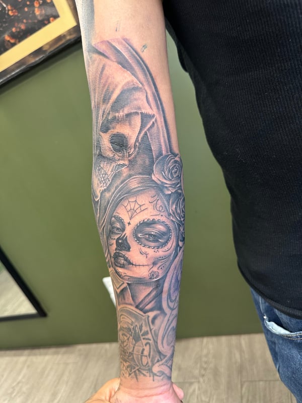 A black and grey Chicano-style tattoo on the forearm featuring a woman's face with skull and rose elements, showcasing detailed shading and realistic features — by Humilde tattoos, los angeles