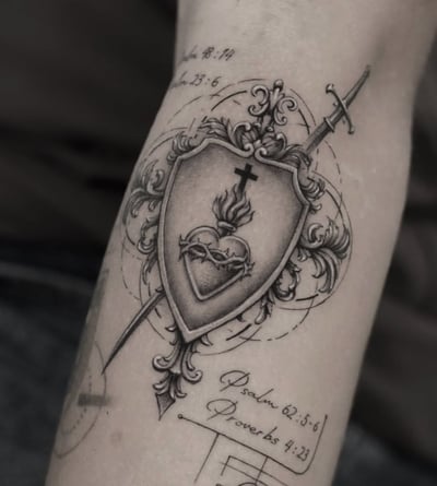 A black and grey ornamental tattoo on the forearm featuring a shield with a flaming heart and cross, pierced by a sword, accompanied by script text — by Jian Liang, los angeles
