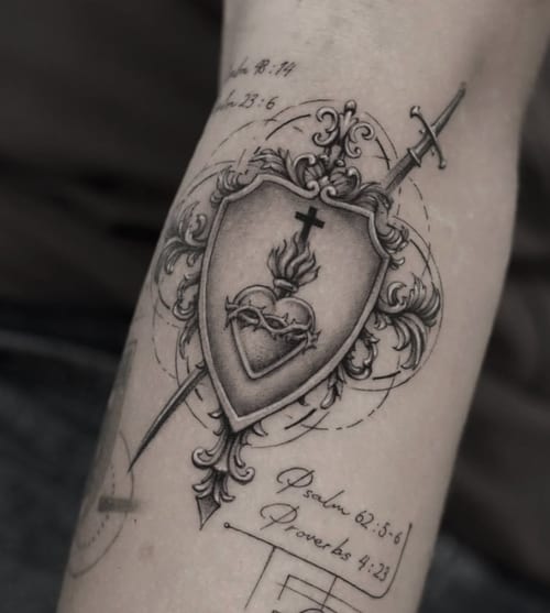 A black and grey ornamental tattoo on the forearm featuring a shield with a flaming heart and cross, pierced by a sword, accompanied by script text — by Jian Liang, los angeles