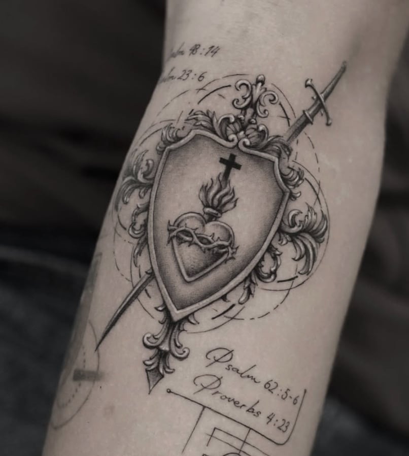 A black and grey ornamental tattoo on the forearm featuring a shield with a flaming heart and cross, pierced by a sword, accompanied by script text — by Jian Liang, los angeles