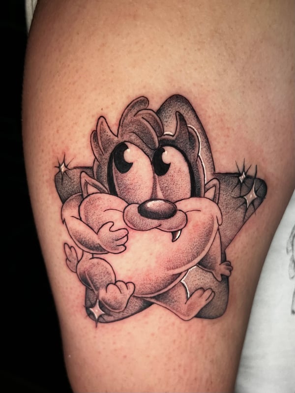 A black and grey cartoon-style tattoo of a character resembling a cartoon animal on the upper arm, featuring bold outlines and shading — by Josue Bravo, los angeles