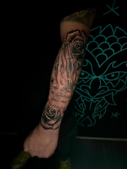 A black and grey realistic tattoo on the forearm featuring praying hands, roses, and script text — by Josue Bravo, los angeles