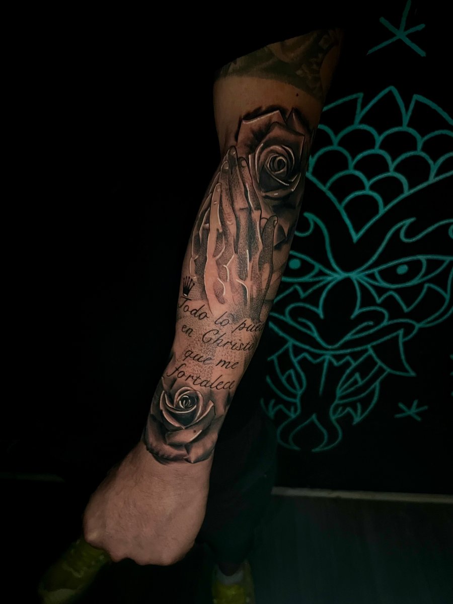 A black and grey realistic tattoo on the forearm featuring praying hands, roses, and script text — by Josue Bravo, los angeles