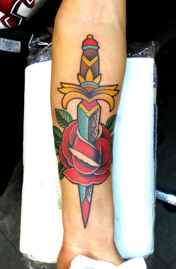 A bold, colorful traditional tattoo of a dagger through a rose on the forearm — by Samuel Connelly, san diego, ca