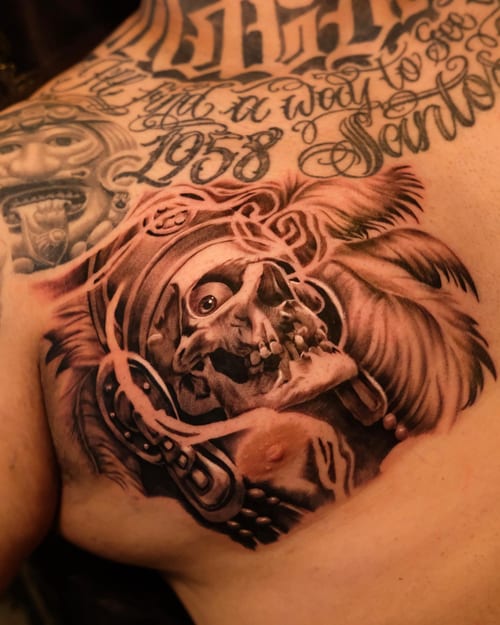A black and grey skull with a headdress tattoo on the chest, featuring ornate script lettering in Chicano style — by Johnny Ayala, los angeles