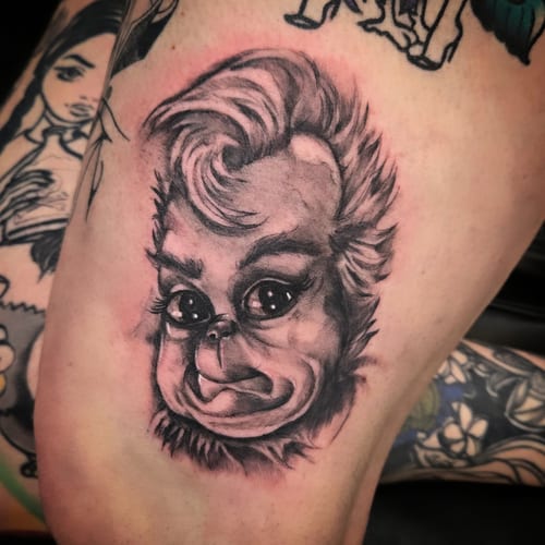 A black and grey illustrative tattoo of a stylized face on the thigh, featuring exaggerated features and shading — by Dretty, los angeles