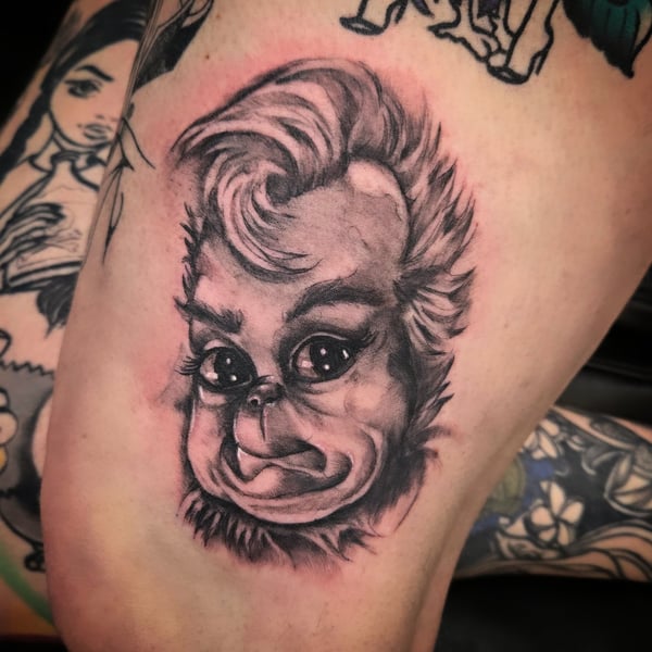 A black and grey illustrative tattoo of a stylized face on the thigh, featuring exaggerated features and shading — by Dretty, long beach, ca
