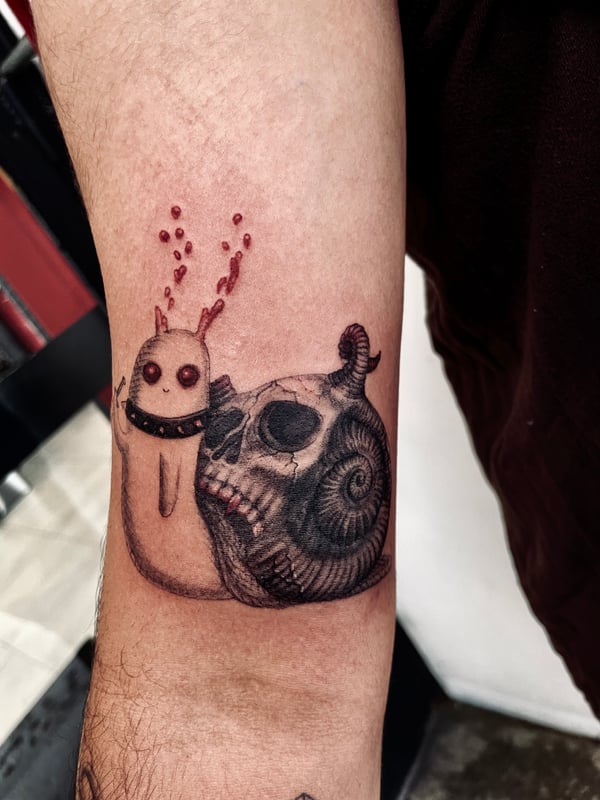 A mixed color and black and grey illustrative tattoo on the forearm, featuring a whimsical creature next to a skull with a snake wrapped around it — by zitronxus, los angeles