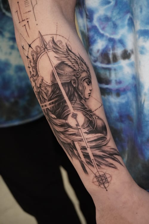 A detailed black and grey tattoo of a mythological warrior woman on the forearm, featuring realistic shading and intricate illustrative elements — by Jian Liang, los angeles