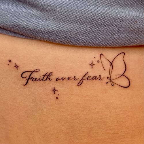 A delicate fine line tattoo on the rib featuring the script 'faith over fear' with a minimalist butterfly design — by NJ TATTOOZ, los angeles