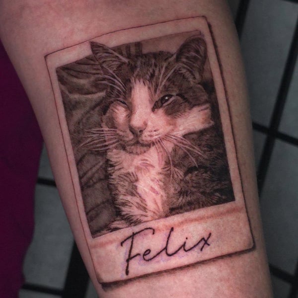 A realistic black and grey cat portrait tattoo on the forearm, styled as a Polaroid photo with the name 'Felix' written below — by Miguel ANGELO, temecula, ca
