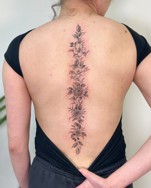 A delicate fine line floral tattoo running vertically along the spine, featuring black and grey ink with intricate flower details — by Melanie Benoit, los angeles