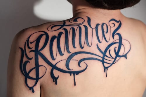 A bold black script lettering tattoo on the back, featuring intricate swirls and flourishes — by Johnny Ayala, los angeles