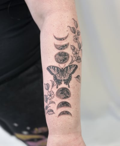 A delicate black and grey tattoo on the forearm featuring a butterfly surrounded by moon phases and floral elements in fine line style — by Melanie Benoit, los angeles