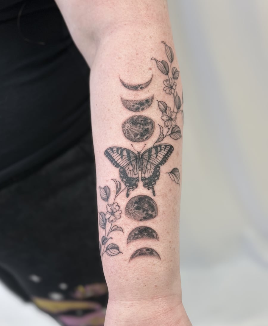 A delicate black and grey tattoo on the forearm featuring a butterfly surrounded by moon phases and floral elements in fine line style — by Melanie Benoit, los angeles