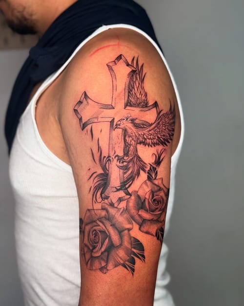 A bold black and grey tattoo featuring a cross with an eagle and roses on the upper arm — by zealous, los angeles