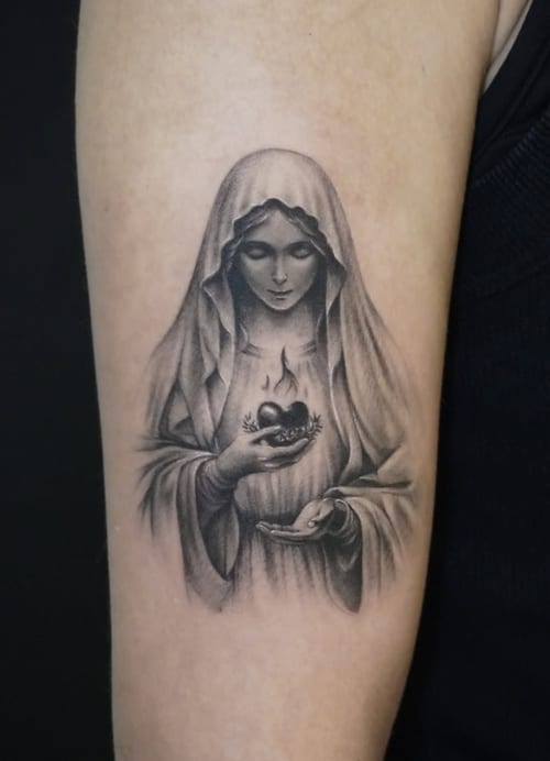 A black and grey realism tattoo of the Virgin Mary holding a flaming heart on the upper arm, with detailed shading and soft lines — by Jian Liang, los angeles