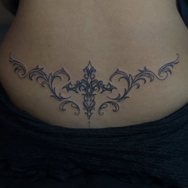 A delicate ornamental cross tattoo on the lower back in fine line style — by Maddie Ramirez, palm desert, ca