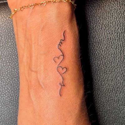 A delicate black ink tattoo on the wrist featuring a fine line heart and script design — by NJ TATTOOZ, los angeles
