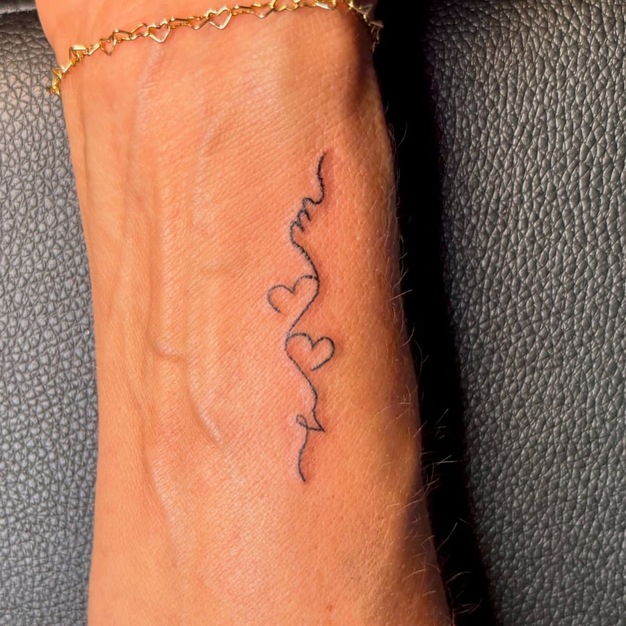A delicate black ink tattoo on the wrist featuring a fine line heart and script design — by NJ TATTOOZ, los angeles
