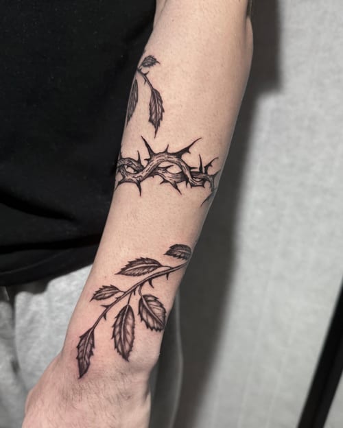 A black and grey fine line tattoo of a thorny vine and leaves wrapping around the forearm with detailed linework — by Shelby Bianco, los angeles