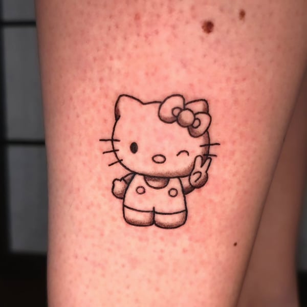 A whimsical black and grey Hello Kitty tattoo on the leg, featuring the iconic cartoon cat in a playful pose — by Miguel ANGELO, temecula, ca
