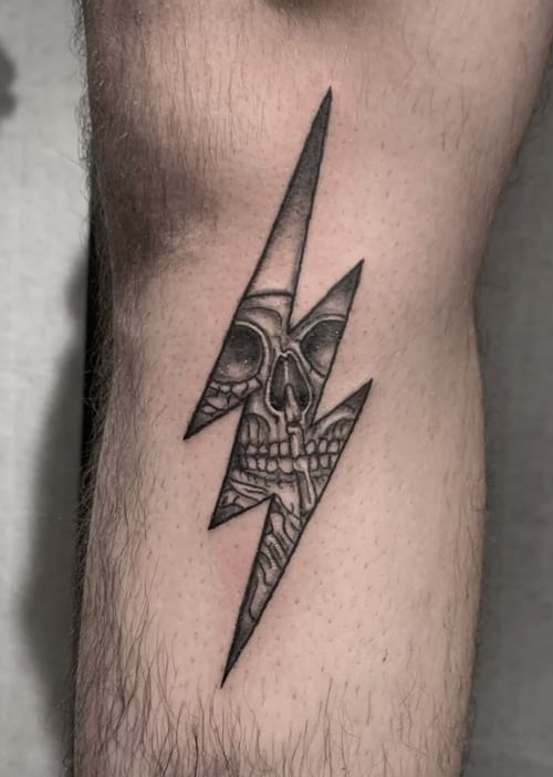 A black and grey tattoo of a skull within a lightning bolt on the forearm, featuring bold linework and shading — by Shelby Bianco, los angeles