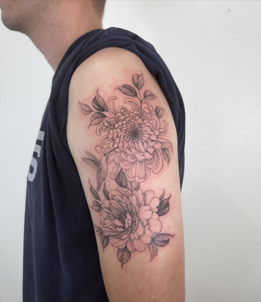 A delicate black and grey tattoo of flowers and a hummingbird on the upper arm, featuring clean lines and intricate floral details — by Melanie Benoit, los angeles