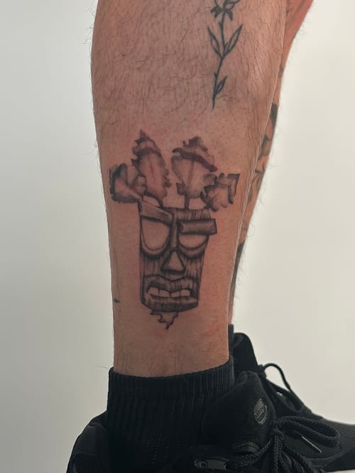 A black and grey tribal-style face tattoo on the calf, featuring bold lines and abstract shapes — by Brian Kies, los angeles