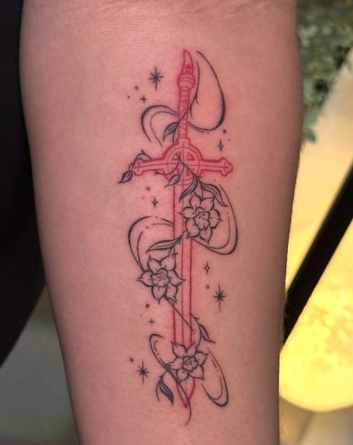 A fine line tattoo on the forearm featuring a sword intertwined with flowers and crescent moons, using a mix of black and red ink — by NJ TATTOOZ, los angeles