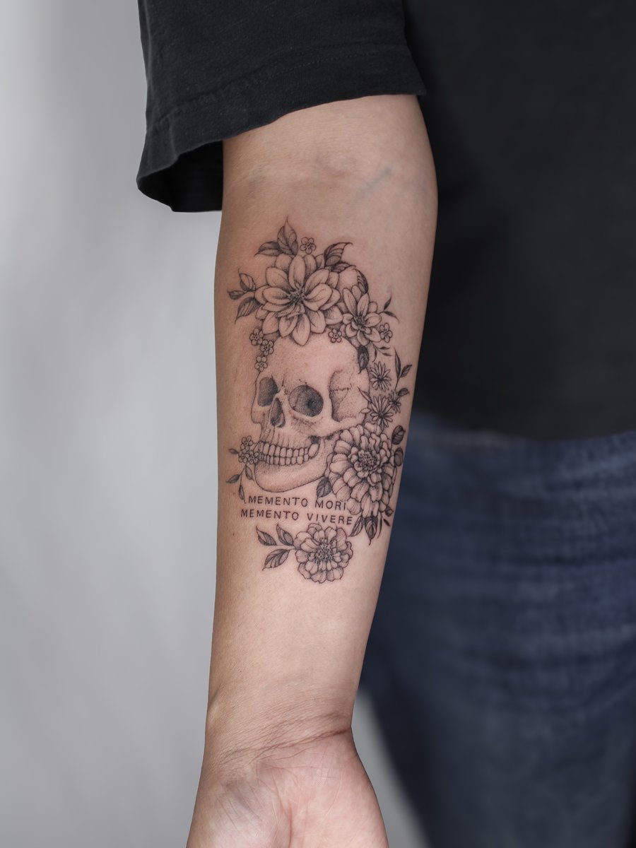 A black and grey skull with flowers and script on the forearm, featuring fine line details and delicate shading — by Melanie Benoit, los angeles