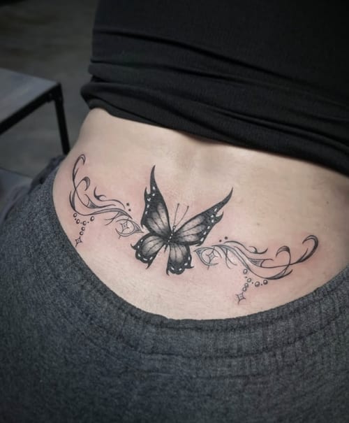 A black and grey butterfly tattoo on the lower back, featuring ornamental floral elements and delicate linework — by Shelby Bianco, los angeles
