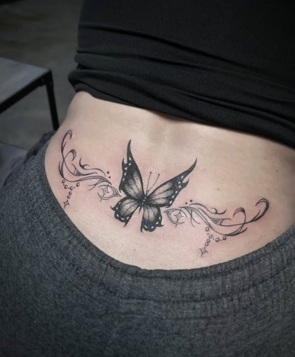 A black and grey butterfly tattoo on the lower back, featuring ornamental floral elements and delicate linework — by Shelby Bianco, ventura, ca