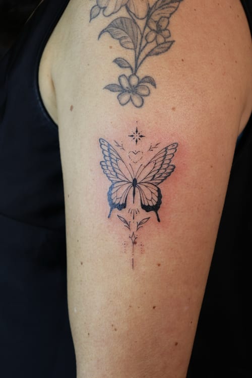 A fine line black and grey butterfly tattoo on the upper arm, featuring delicate floral accents and dot shading — by gloria.shi.ink, los angeles
