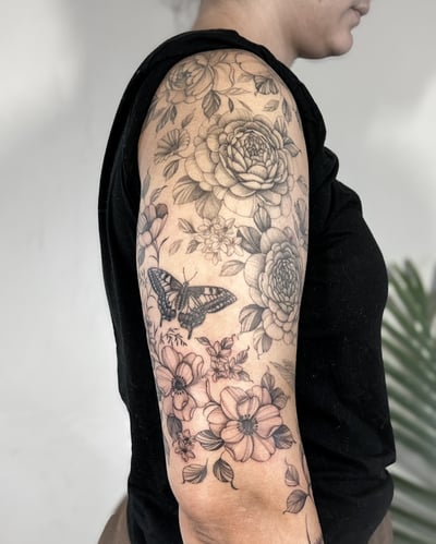 A delicate black and grey floral tattoo with a butterfly on the upper arm, featuring detailed flowers and leaves — by Melanie Benoit, los angeles