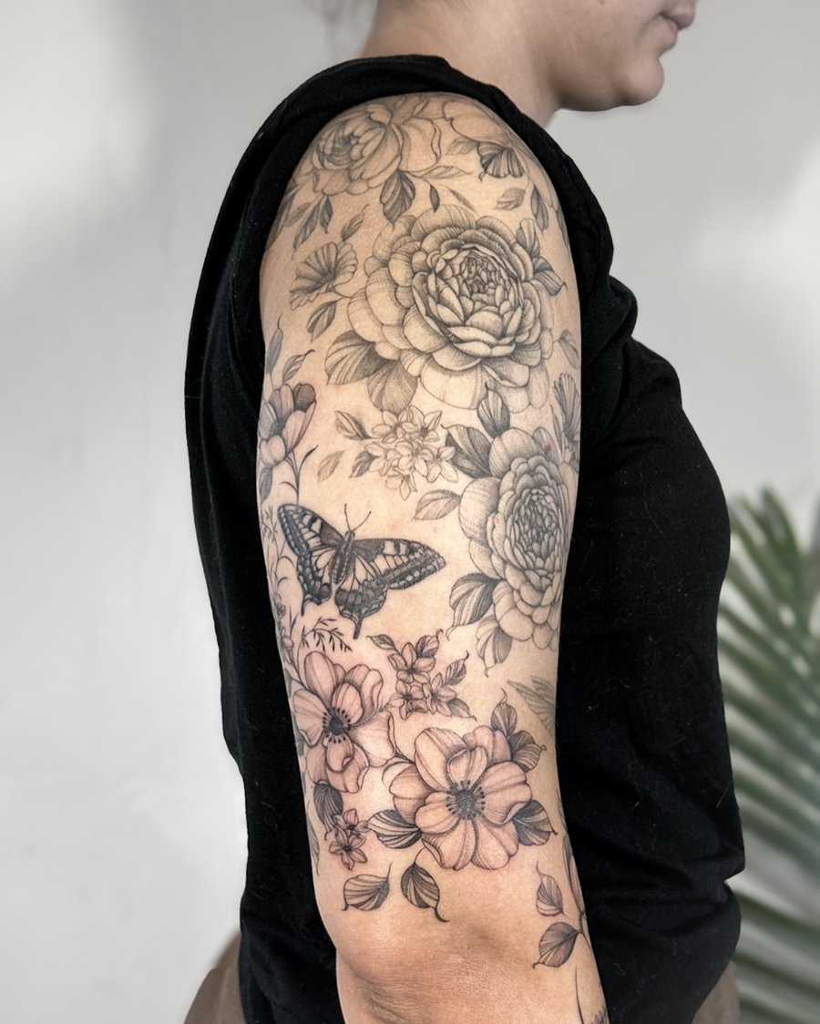 A delicate black and grey floral tattoo with a butterfly on the upper arm, featuring detailed flowers and leaves — by Melanie Benoit, los angeles