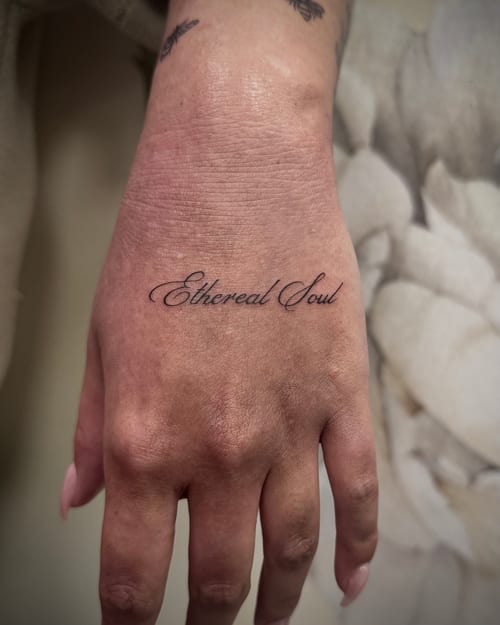 A minimalist black and grey script tattoo on the hand reading 'Ethereal Soul' in elegant cursive lettering — by emily, los angeles