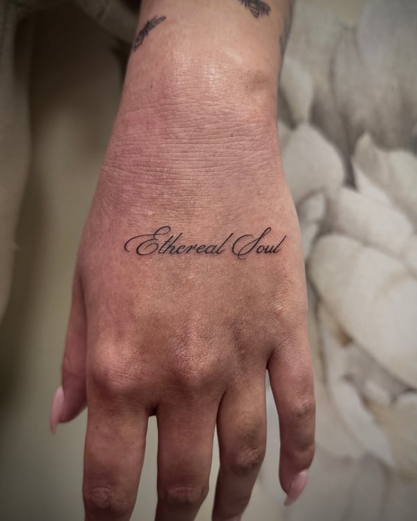A minimalist black and grey script tattoo on the hand reading 'Ethereal Soul' in elegant cursive lettering — by emily, rahway