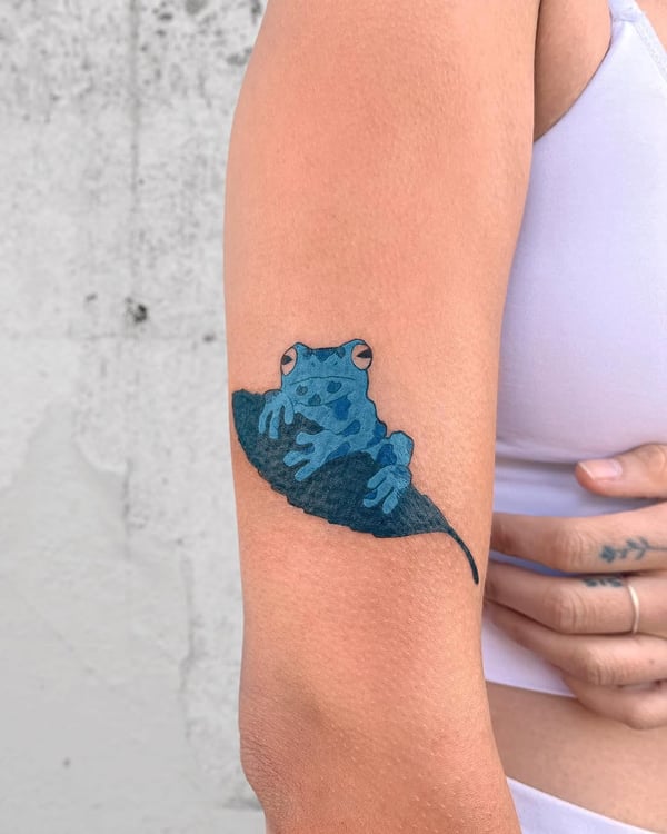 A whimsical and colorful tattoo of a blue frog sitting on a leaf, located on the upper arm — by Dylan Aiello, los angeles