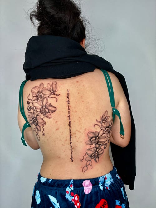 A black and grey fine line tattoo on the back featuring detailed flowers and script running vertically between them — by Edgar Armas Rodriguez, los angeles